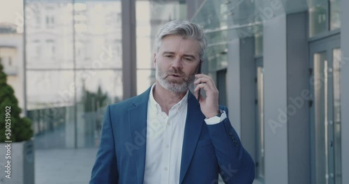 Business Person Holding Smartphone On Go During Break. Senior Businessman Talking Mobile Walking On Street. Mature Guy Call To Colleague Near Office Building. Business and Lifestyle Concept 