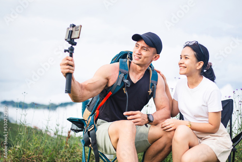 Asian tourist couple with backpack taking selfie photo on vacation trip. Happy traveler man and woman shooting video with selfie stick on mountain and fog background. Blogger and vlog concept. © Pun