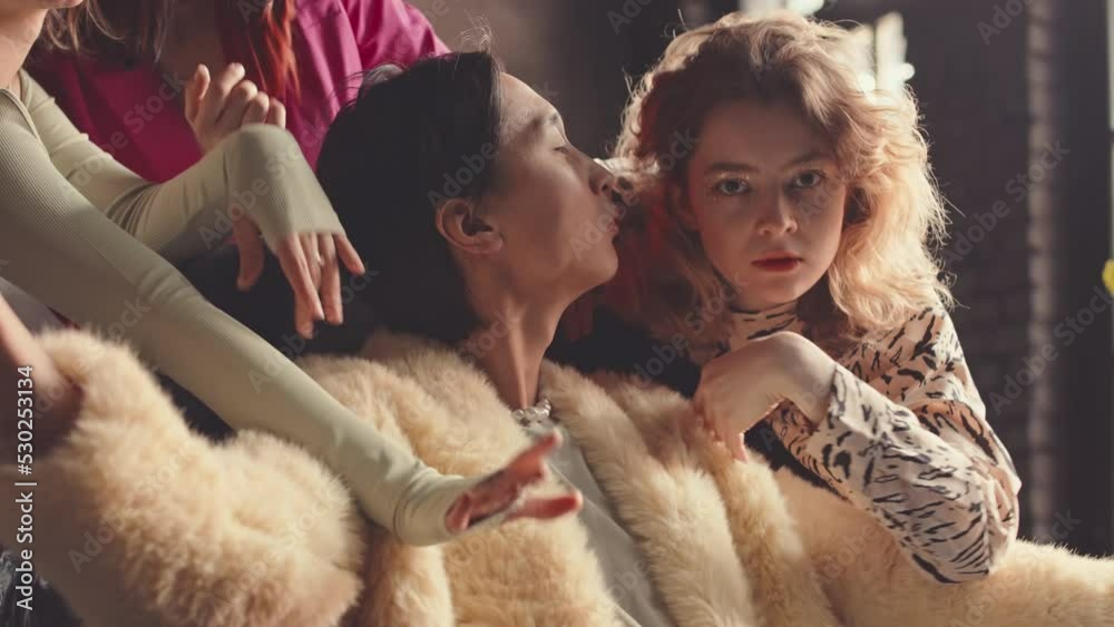 Slowmo of confident Asian guy in camel fur coat looking at camera pompously while sitting in chair among three beautiful fancy girls dancing vogue to him Slowmo of confident Asian guy in camel fur coat looking at camera pompously while sitting in chair among three beautiful fancy girls dancing vogue to him