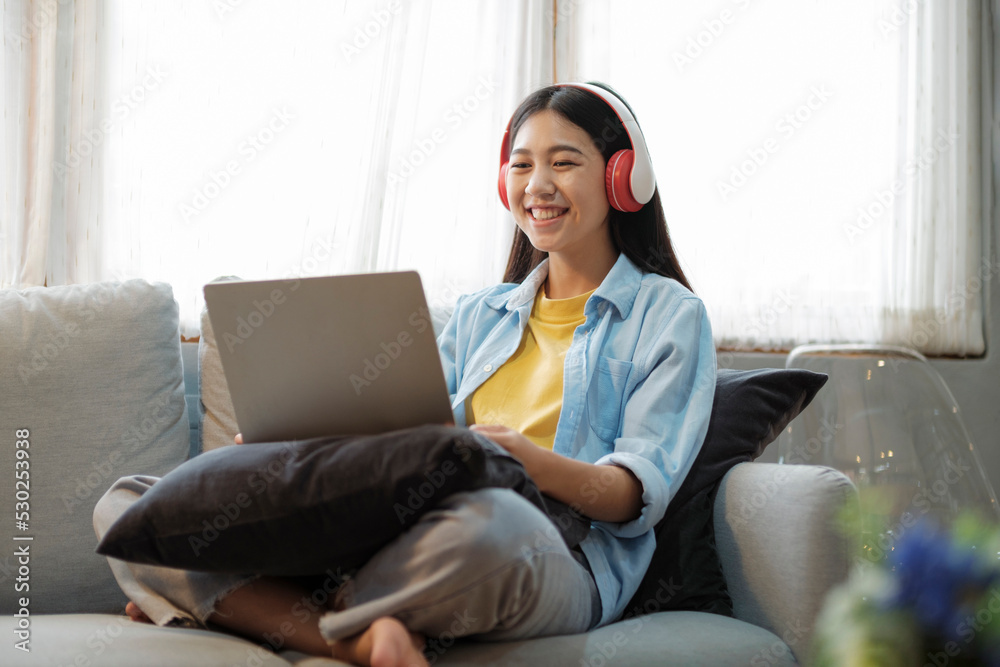 Naklejka premium Young asian woman smiling and listening to music using laptop while sitting on couch at home.