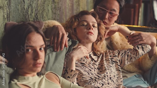 Two attractive girls and pompous Asian guy in fur coat looking at camera and making seductive moves sitting on sofa in authentic studio photo