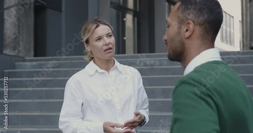 Elegant Colleagues Discussing Project Standing on Street. Multiracial Young Man and Senior Woman Talking with Each Other. Urban Background 