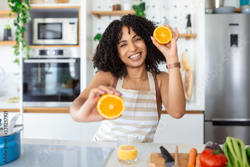 Photo of cute African American woman smiling and holding two orange parts while cooking vegetable salad in kitchen interior at home