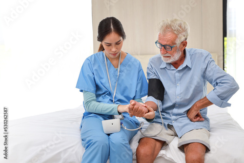 Happy caregiver checking blood pressure monitor and heart rate senior elderly man patient on bed at retirement house. Asian smiling nurse taking good help care and support of elder patient at house