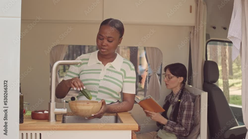 Waist up shot of two young pretty multiethnic girlfriends spending summer weekends outdoors in cozy house on wheels. One of girls washing vegetables for lunch while other reading book Waist up shot of two young pretty multiethnic girlfriends spending summer weekends outdoors in cozy house on wheels. One of girls washing vegetables for lunch while other reading book