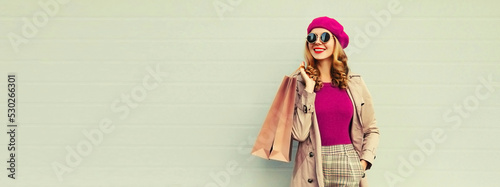 Autumn portrait of beautiful happy smiling woman with shopping bags wearing beige coat, pink french beret on gray background photo