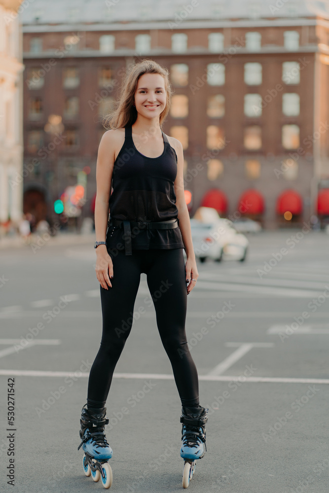 Positive young female model in sportsclothes rides on blades enjoys leisure activities poses at urban place against blurred background stands in full length. Active lifestyle and rollerblading concept