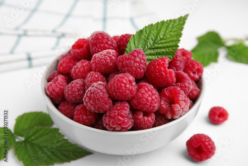 Bowl of fresh ripe raspberries with green leaves on white table