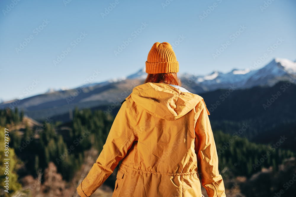 Woman standing in the fall back up her hands up in the mountains nature hike and happiness in yellow cape with red hair full-length standing in front of the trees and mountains in the sunset, freedom