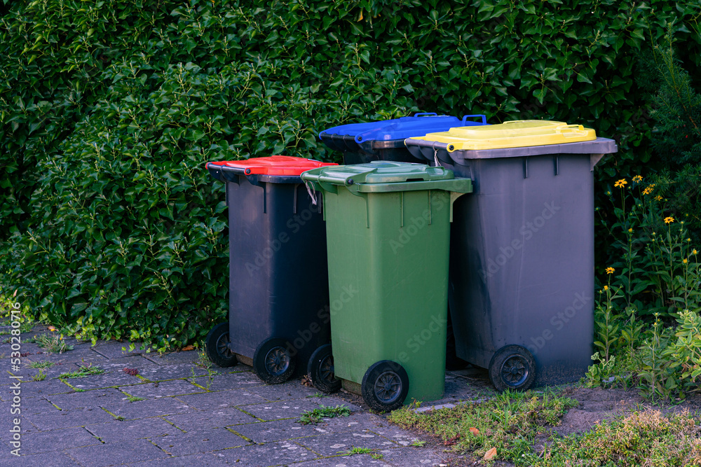 Group of plastic trash cans whit multicolored lid. Green bushes in the background. Group of plastic trash cans whit multicolored lid. Green bushes in the background.