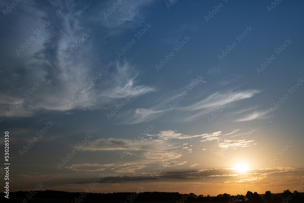 The sun goes down on the horizon. The sky is blue with white clouds. The sky glows red on the horizon. The outlines of trees can be seen at the bottom.