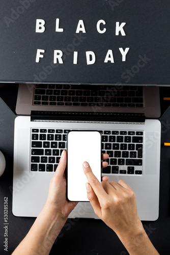 Composition of hand using smartphone with black friday text and laptop on gray background