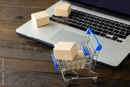 Composition of shopping cart with boxes and laptop on wooden background