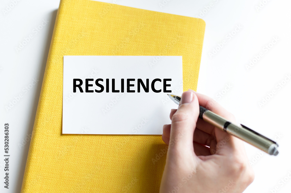 RESILIENCE text on a white card against the background of a yellow notebook next to the hand of the businessman with a pen writes the text RESILIENCE text on a white card against the background of a yellow notebook next to the hand of the businessman with a pen writes the text