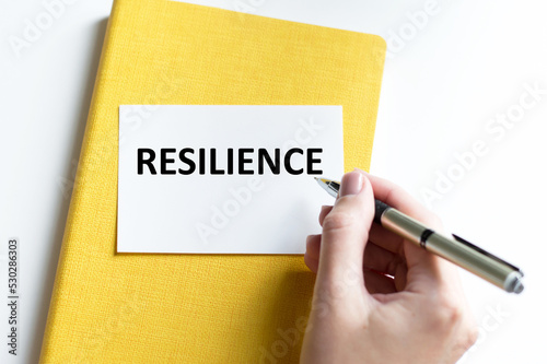 RESILIENCE text on a white card against the background of a yellow notebook next to the hand of the businessman with a pen writes the text