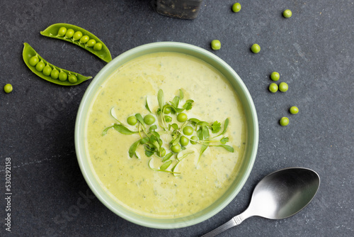 Horizontal image of bowl of pea soup with garnish, and peas and pea pods on slate, copy space