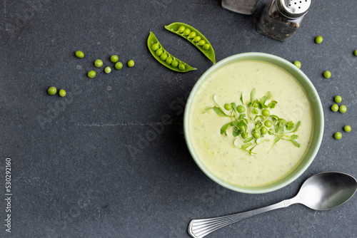Horizontal image of bowl of pea soup with garnish, and peas and pea pods on slate, copy space