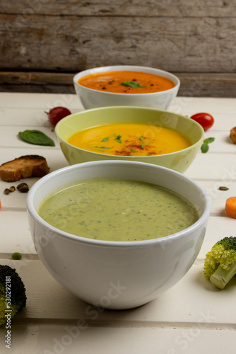 Vertical image of bowls of carrot, broccoli and tomato soups on white wood, with copy space