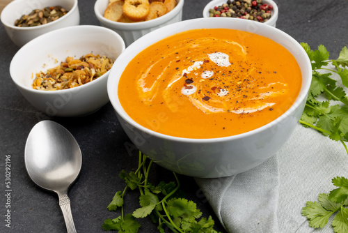 Horizontal image of bowl of tomato soup, with toast, seeds, leaves and peppercorns on slate
