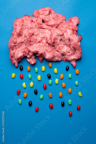 Vertical image of cloud of homemade pink candy floss raining jelly beans, on blue with copy space