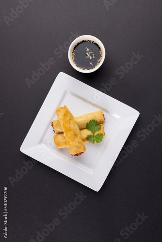 Overhead view of asian spring rolls on white plate and soy sauce on grey background