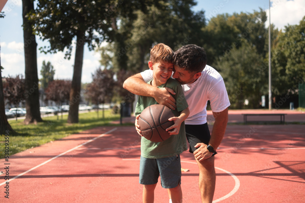 Smiling father hugging his son on basketball court. They wear casual clothes, boy holding a basketball