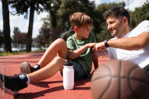 Fototapeta Naklejka Na Ścianę i Meble -  Father and son lying on basketball court, giving fist Bump to each other. They enjoy the day after had basketball training. Boy holding plastic water bottle 