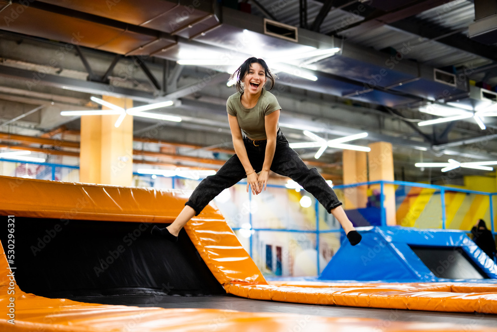 Naklejka premium Pretty girl jumping on colorful trampoline at playground park, posing and laughing. Beautiful female teenager happy during active entertaiments indoor