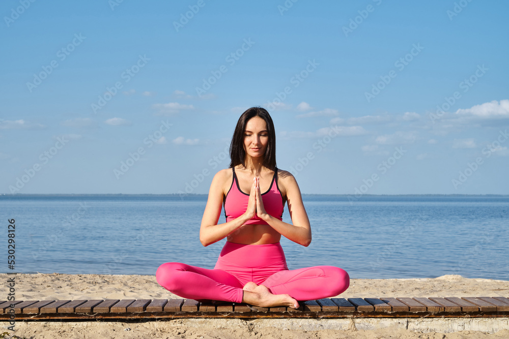 Women in sportswear sitting in a lotus position and meditating during yoga classes on the seashore. Practicing yoga lesson, breathing, meditation, doing Ardha Padmasana exercises. Wellness concept