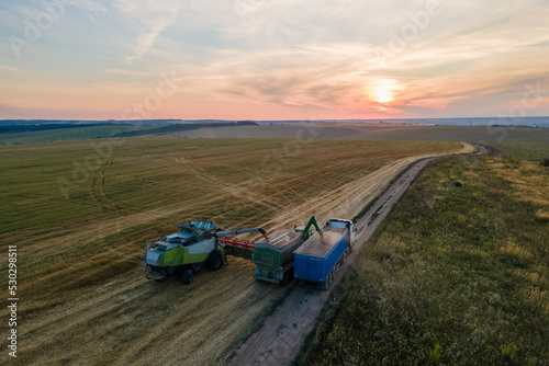 Aerial view of combine harvester unloading grain in cargo trailer working during harvesting season on large ripe wheat field. Agriculture and transportation of raw farm products concept