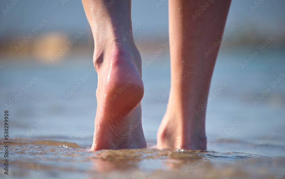 Close up of female feet walking barefoot on white grainy sand of golden beach on blue ocean water background