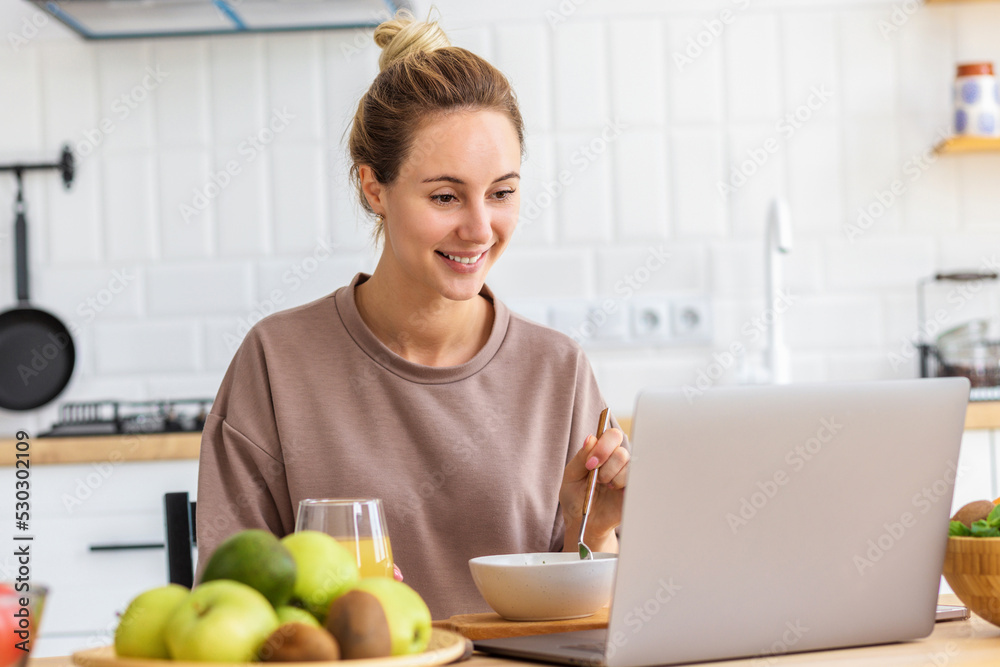 Happy young Caucasian woman healthy food breakfast and chatting online using laptop sitting at home in the kitchen Happy young Caucasian woman healthy food breakfast and chatting online using laptop sitting at home in the kitchen