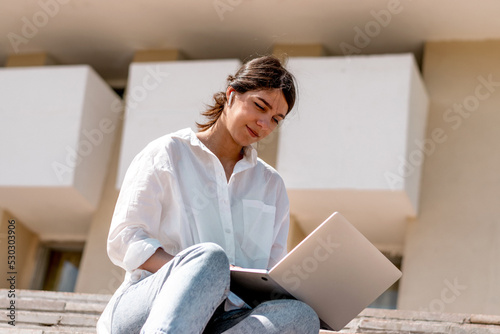 A young dark-haired woman in a white shirt and jeans uses a laptop sitting on the steps on a city street on a summer day.Remote work  freelance online education.