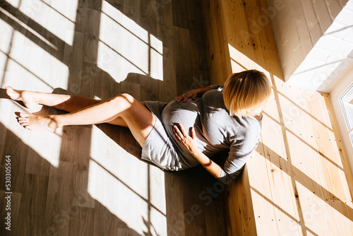 Pregnant blonde lies on the floor in a gray dress from the window rays from the sun