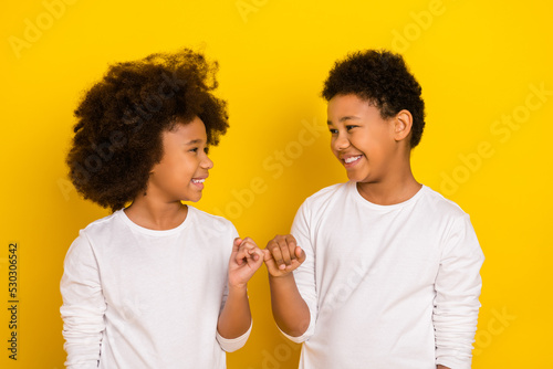 Photo of sweet friendly schoolboy schoolgirl wear white shirts holding small fingers isolated yellow color background photo