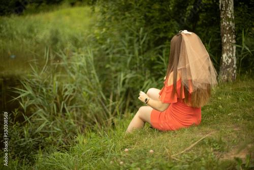 Girl on shore of lake in summer. Maiden in orange clothes. Bride in dress. Holiday in park.