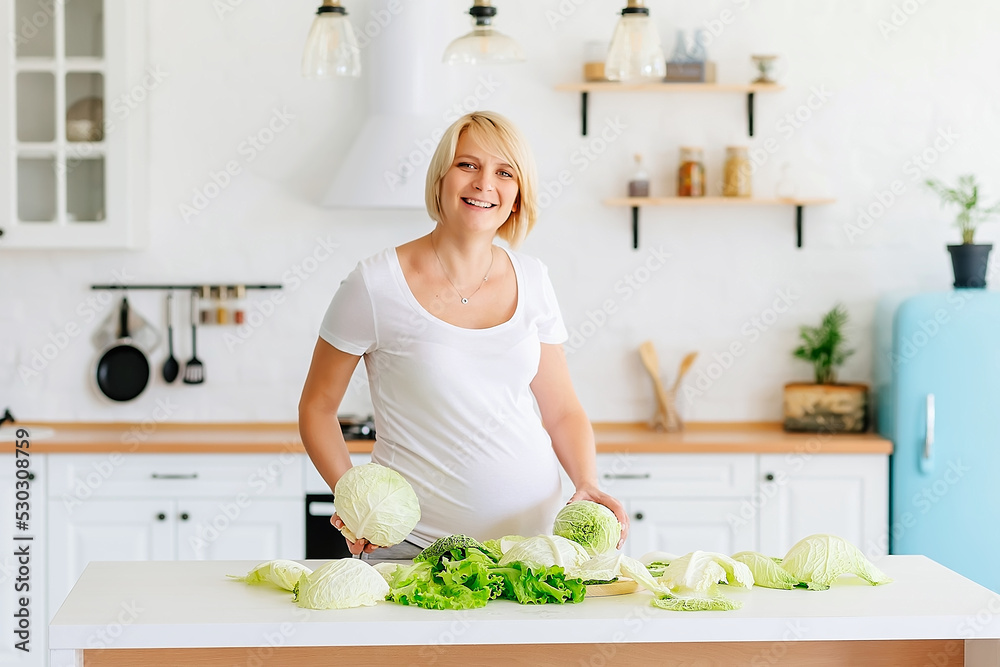 Beautiful blonde girl pregnant blonde in the kitchen in Scandinavian style at the table parses green cabbage