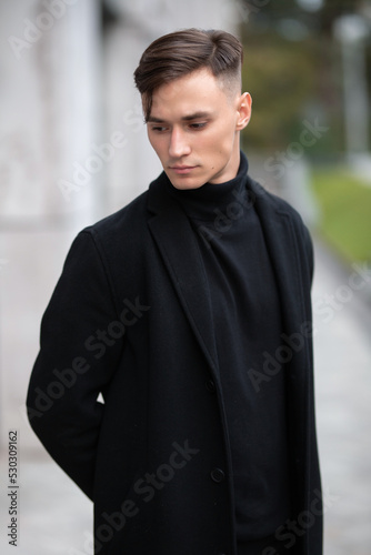 Fashion portrait of a young guy in black clothes of model appearance on the street among skyscrapers and alleys