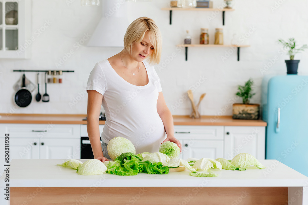 Beautiful blonde girl pregnant blonde in the kitchen in Scandinavian style at the table parses green cabbage 