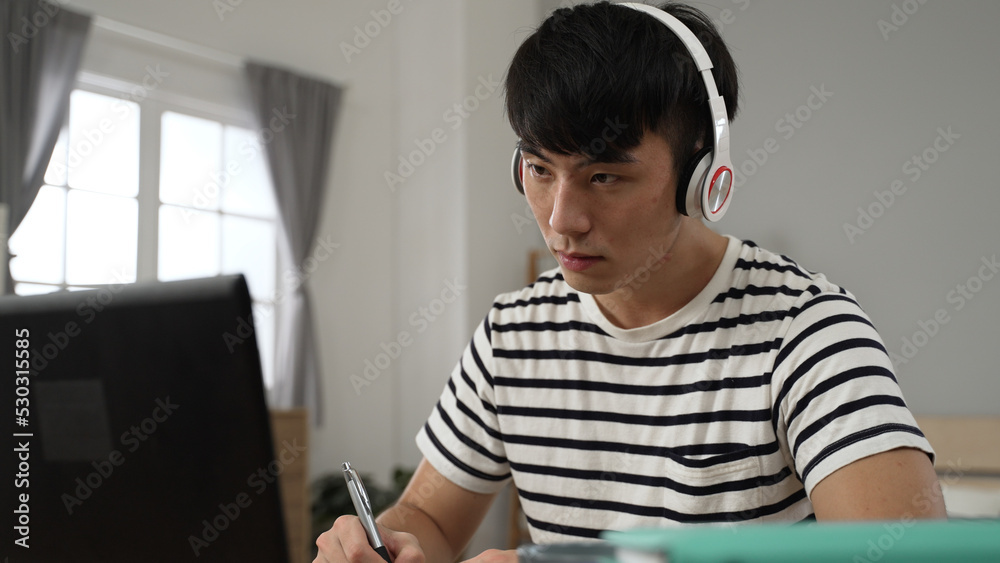 smiling asian student learning from home with headphones is nodding head to show understanding and taking notes while taking virtual lesson on laptop in bedroom smiling asian student learning from home with headphones is nodding head to show understanding and taking notes while taking virtual lesson on laptop in bedroom