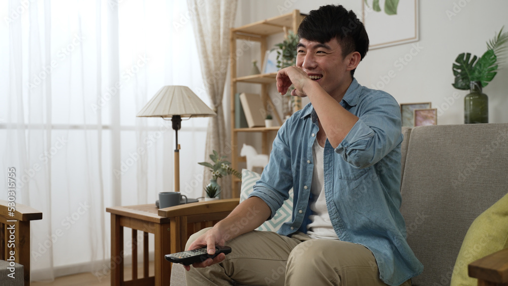 leisure Korean young man with tv controller is laughing and slapping on his lap while watching a funny comedy show in the living room at home during daytime