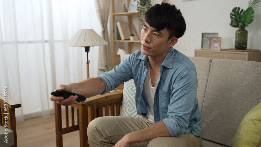 asian Korean guy shaking head and feeling dissatisfied while channel surfing with a remote control on the sofa in the living room at home asian Korean guy shaking head and feeling dissatisfied while channel surfing with a remote control on the sofa in the living room at home