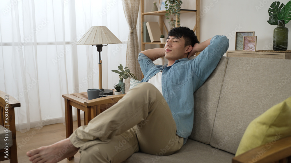 leisure Asian guy is stretching body and placing arms behind head to take a nap with folded legs on comfortable sofa in the living room at home during daytime