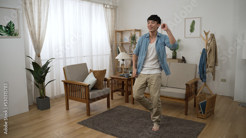 full length view of an energetic asian young guy having fun dancing to music with barefoot in a modern home living room interior with daylight.