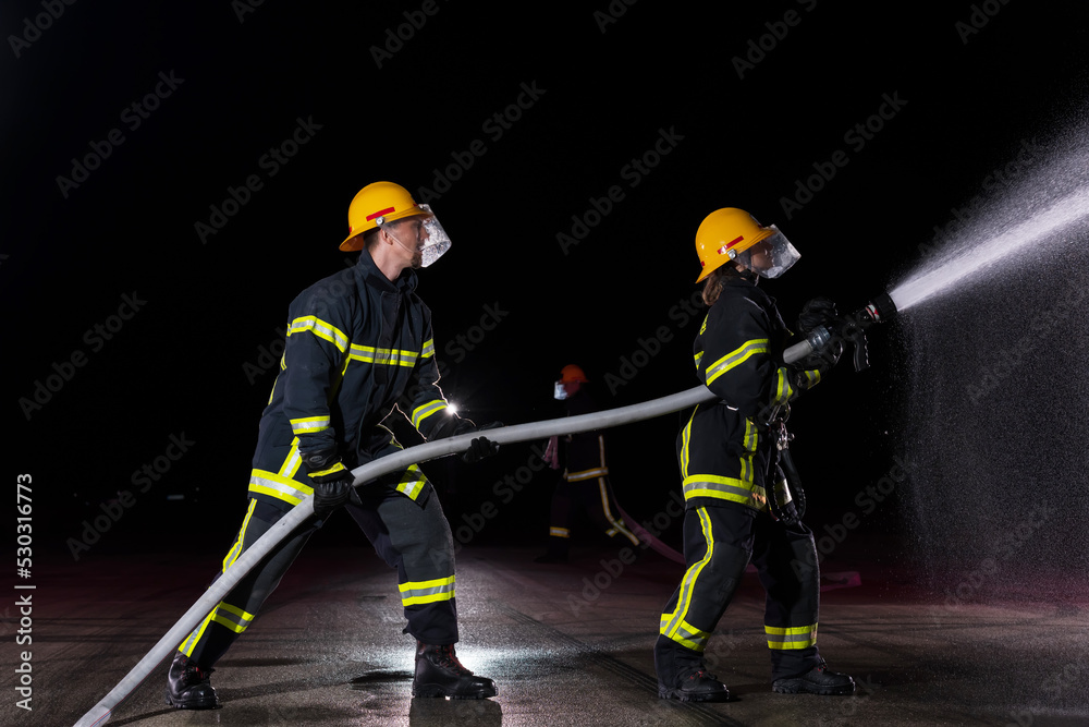 Fototapeta premium Firefighters using a water hose to eliminate a fire hazard. Team of female and male firemen in dangerous rescue mission. 