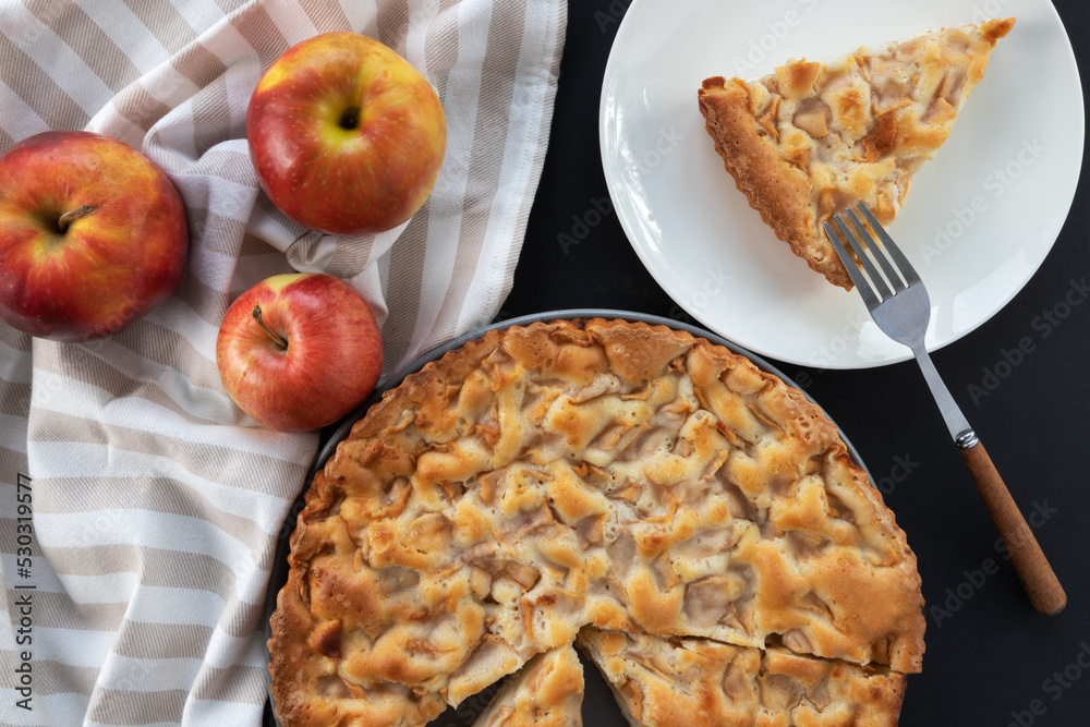 Homemade apple pie on the table in a gray plate with red apples and a kitchen towel. A piece of cake in a white plate with fork. Top view banner. Flat background with pastries Homemade apple pie on the table in a gray plate with red apples and a kitchen towel. A piece of cake in a white plate with fork. Top view banner. Flat background with pastries