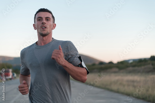 Attractive fit man running fast along countryside road at sunset light, doing jogging workout outdoors