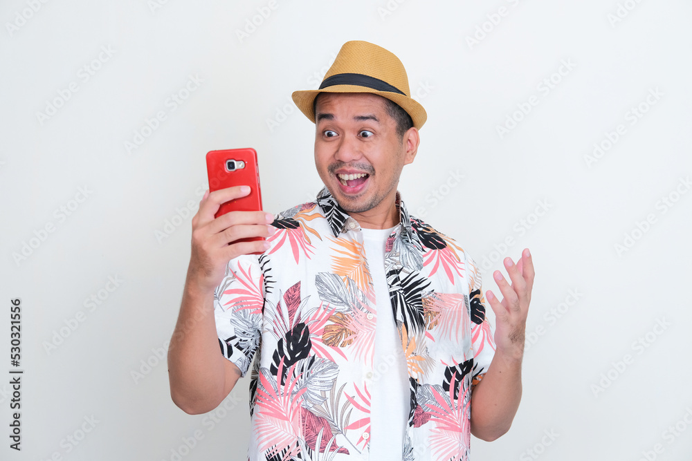 Asian man wearing beach shirt showing excited when looking to his handphone Asian man wearing beach shirt showing excited when looking to his handphone
