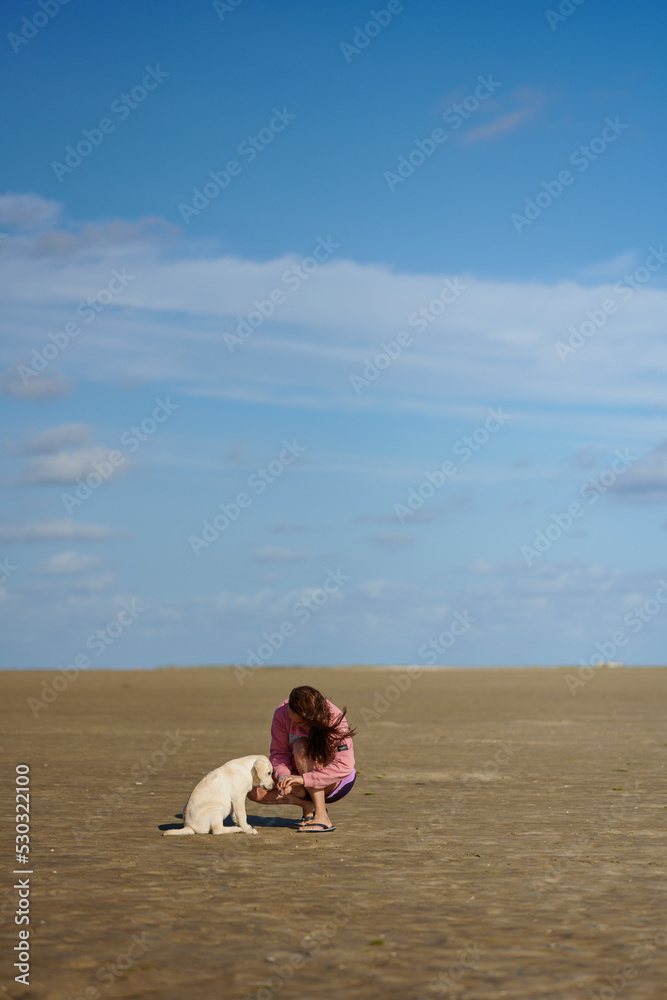 Attractive middle-aged woman feeding her puppy at the beach kneeling on the sand with copy space above