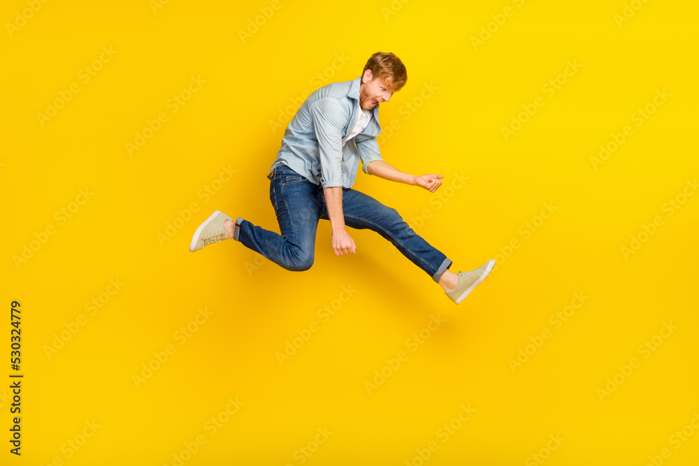 Full size photo of handsome guy ginger hairdo blue shirt jeans sneakers jump play imaginary guitar isolated on yellow color background